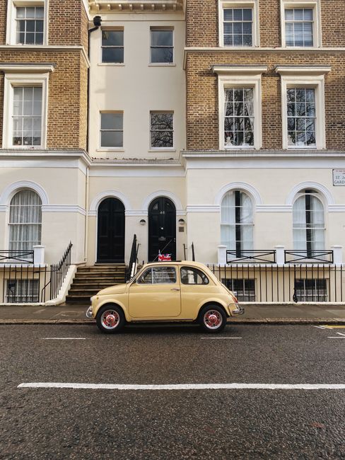A vintage beige Fiat 500 with red wheels is parked on the street in front of a set of stone steps leading up to the entrance of a multi-story residential building. The building features a combination of brick and painted exterior walls, with a white façade below and brown brickwork above. The ground-floor windows are arched and have white frames, with some covered by curtains, and a black front door is situated between the steps. A Union Jack flag is attached to the front of the car. The street surface is dark asphalt with visible white road markings, and a metal railing surrounds the stairs, leading to black railings on either side of the entrance. The scene is lit with natural daylight, capturing a typical urban setting that could relate to private waste collection or alternative rubbish management in a city environment. The image is clear and detailed, with a focus on the vehicle and building façade, often seen in contexts of residential waste clearance or off-site rubbish removal services provided by companies like Waste Disposal Bayswater.