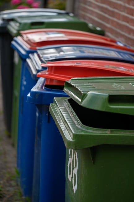 A row of large, rectangular wheelie bins positioned along a sidewalk adjacent to a brick wall. The bins are made of durable plastic with textured lids, and they are painted in distinct colors including green, blue, and red. The green bin, closest to the camera, is slightly tilted forward, revealing its ribbed lid with a hinged opening. The blue bins have smooth lids with some visible white markings or labels. All bins are fitted with wheels at the base, and they are arranged in a neat, linear formation, possibly awaiting collection or disposal. The scene is outdoors, with natural daylight illuminating the bins, highlighting their textures and colors, and the background trees and foliage are slightly blurred. The setup suggests an area designated for waste management, with Waste Disposal Bayswater specializing in alternative waste handling and rubbish removal services for local property cleaning or on-site clearance needs.