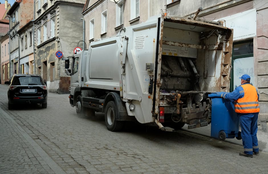 A worker wearing a blue uniform and a high-visibility orange vest is positioned at the rear of a large white rubbish collection truck on a narrow urban street, actively engaged in waste collection. The worker is holding a blue wheeled rubbish bin and appears to be about to empty it into the truck’s open hopper, which is lined with rusted, brownish metal edges and has a mechanical lifting mechanism visible inside. The truck’s rear section shows signs of dirt and use, with various mechanical components and rusted areas. In the background, the street is lined with older, multi-storey buildings featuring neutral-colored facades, some with peeling paint and worn plaster, and a black car is parked on the left side of the street. The scene is illuminated by natural daylight, providing clear visibility of the urban environment and the waste collection activity, illustrating a typical private rubbish removal operation within a city setting, supported by Waste Disposal Bayswater’s waste management services.