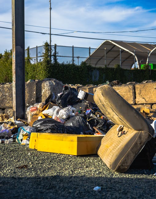 An unmanaged pile of mixed waste materials is situated on a gravel surface, featuring black garbage bags, discarded cardboard, plastic packaging, and crumpled papers. A large, used car tire is leaning against the pile, with its worn tread and weathered sidewall visible, and a bright yellow plastic container is partially buried among the debris. In the background, a stone wall made of irregularly shaped rocks separates the waste from a fenced-off area with shrubbery and tall trees. Overhead, power lines stretch across a clear blue sky, suggesting a semi-urban environment. To the right, part of a structure with a curved, semi-transparent roof or canopy is visible, possibly an outdoor storage or industrial facility. The scene illustrates an example of improper waste disposal, which services like Waste Disposal Bayswater aim to address through alternative rubbish removal solutions, including private collection and on-site clearance, depending on specific waste management needs.