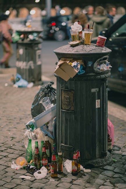 A worker wearing a blue uniform and a high-visibility orange vest is positioned at the rear of a large white rubbish collection truck on a narrow urban street, actively engaged in waste collection. The worker is holding a blue wheeled rubbish bin and appears to be about to empty it into the truck’s open hopper, which is lined with rusted, brownish metal edges and has a mechanical lifting mechanism visible inside. The truck’s rear section shows signs of dirt and use, with various mechanical components and rusted areas. In the background, the street is lined with older, multi-storey buildings featuring neutral-colored facades, some with peeling paint and worn plaster, and a black car is parked on the left side of the street. The scene is illuminated by natural daylight, providing clear visibility of the urban environment and the waste collection activity, illustrating a typical private rubbish removal operation within a city setting, supported by Waste Disposal Bayswater’s waste management services.