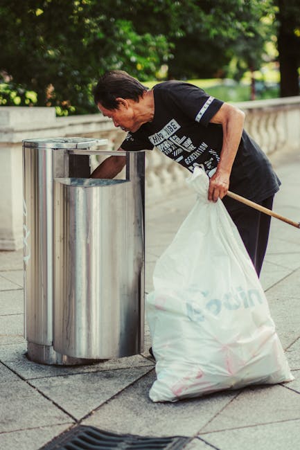 A woman with short dark hair, wearing a black T-shirt with white text on the front, is seen composting litter by depositing rubbish into a large, shiny, stainless steel public litter bin on a paved sidewalk. She is holding a white cloth bag filled with waste in her left hand and using her right hand to reach into the bin, which is placed next to a low stone balustrade. The bin has an open top with a slight rim and is designed for outdoor use, positioned in a green, tree-lined environment with dappled sunlight filtering through the foliage. The scene depicts a typical public space where private waste disposal services, such as those provided by Waste Disposal Bayswater, can facilitate on-site clearance of rubbish, supporting local efforts for effective waste management outside of council collection. The man’s posture and action suggest an organized approach to waste removal, highlighting the importance of proper rubbish handling in urban outdoor areas.