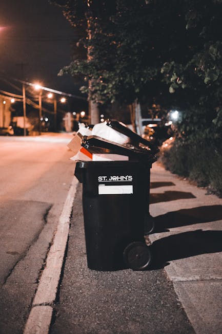 A red clinical waste bin with a hinged lid, positioned on a concrete pavement near a street. The bin has a diamond-shaped hazardous waste symbol with the text 'CLINICAL WASTE' in English and Chinese, indicating it is used for medical or biological waste. The surface of the bin appears slightly scuffed and the wheels at its base are black with a metal framework, allowing for easy mobility. In the background, part of a white waste collection truck and some pedestrians are visible, along with a few street markings such as double yellow lines. The scene suggests an urban environment where waste is being managed through private collection services, and Waste Disposal Bayswater could be involved in the handling or removal of such clinical waste containers. The overall setting is outdoors, with natural ambient lighting highlighting the textures of the plastic surface and the surrounding street elements.